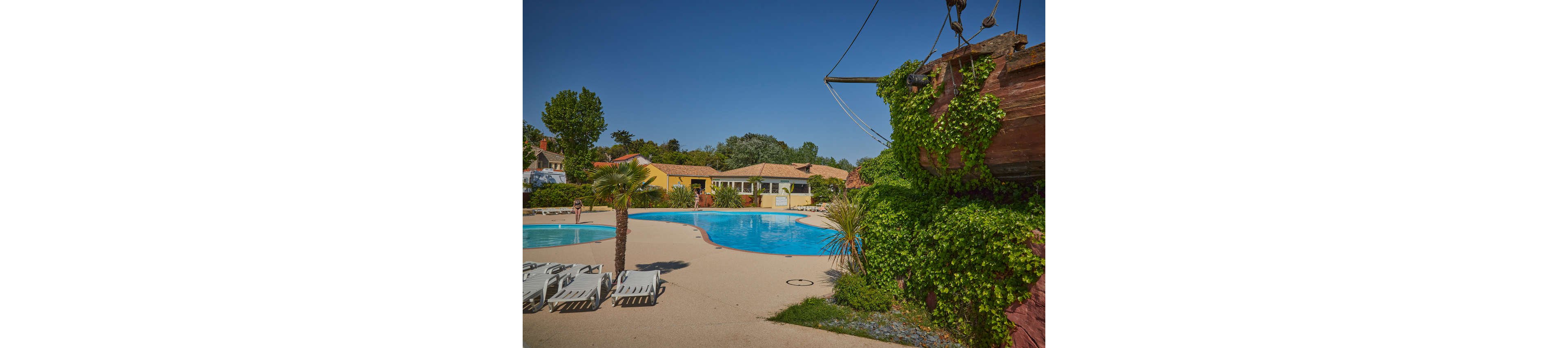 An outdoor resort swimming pool is set under a clear blue sky, surrounded by sun loungers and palm trees. A few people enjoy the water while colorful low-rise buildings form the backdrop. On the right, a structure resembling a wooden ship is covered in lush green ivy, adding a playful, tropical touch. The bright sunlight and clear weather create a relaxed vacation atmosphere.