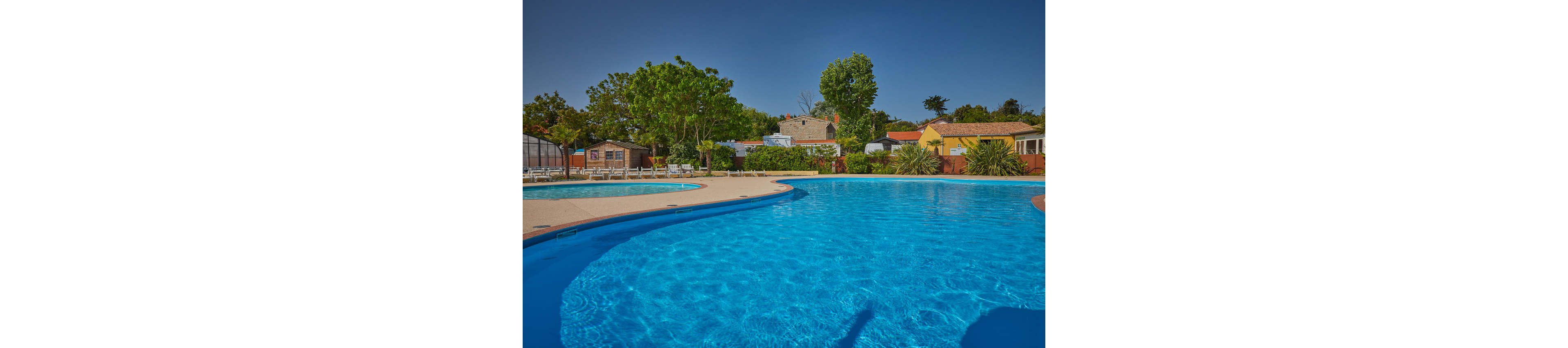 A large, clear blue swimming pool curves through the foreground of an outdoor resort. Sun loungers line the far side of the pool, facing toward the water under a bright blue sky. Behind the pool are small houses and cabins surrounded by green trees and shrubs. The scene suggests a quiet, relaxing vacation setting in warm weather.