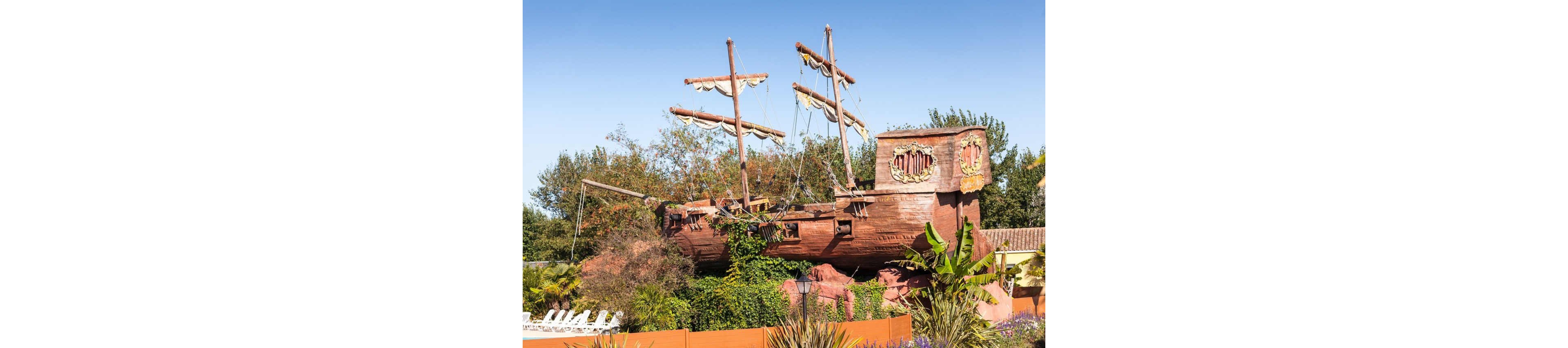 A large, weathered pirate ship structure is displayed outdoors, resembling a classic wooden galleon with broken masts and rigging. The ship appears to be part of an amusement or theme park attraction, surrounded by lush greenery and tropical plants. Decorative details and carved emblems are visible on the ship’s sides, enhancing its fantasy appearance. A clear blue sky and distant trees frame the scene, emphasizing the playful, adventurous atmosphere.