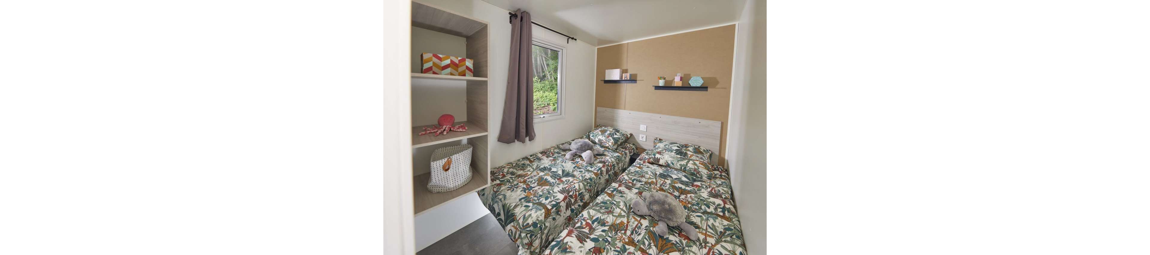 A compact bedroom features two single beds with matching tropical-patterned duvets and plush toys placed on top. The room includes a small window with a dark curtain, allowing natural light and a view of greenery outside. Open shelves on the left hold decorative boxes and a woven basket, while wall-mounted shelves above the headboards display small decor items. Neutral walls and light wood furniture create a calm, modern atmosphere suitable for a child or guest room.