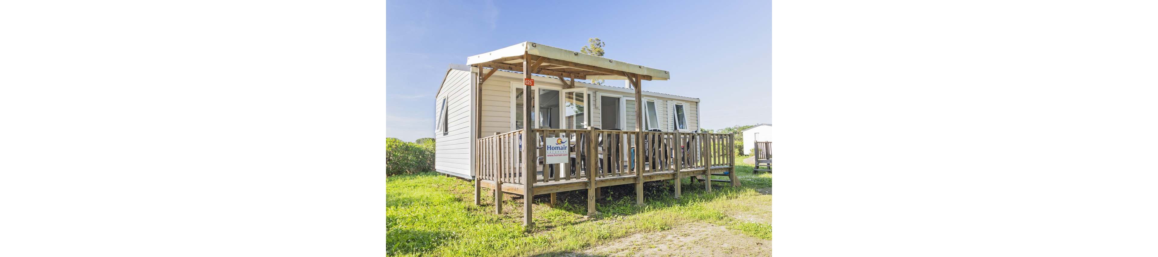 A light-colored mobile home with a wooden terrace stands on a grassy plot under a clear blue sky. The structure features multiple windows and a covered porch area with railings. A small sign on the terrace reads "Homair" and a red number plate shows "825". The surrounding area appears to be part of a campground or holiday park with other similar units in the background.