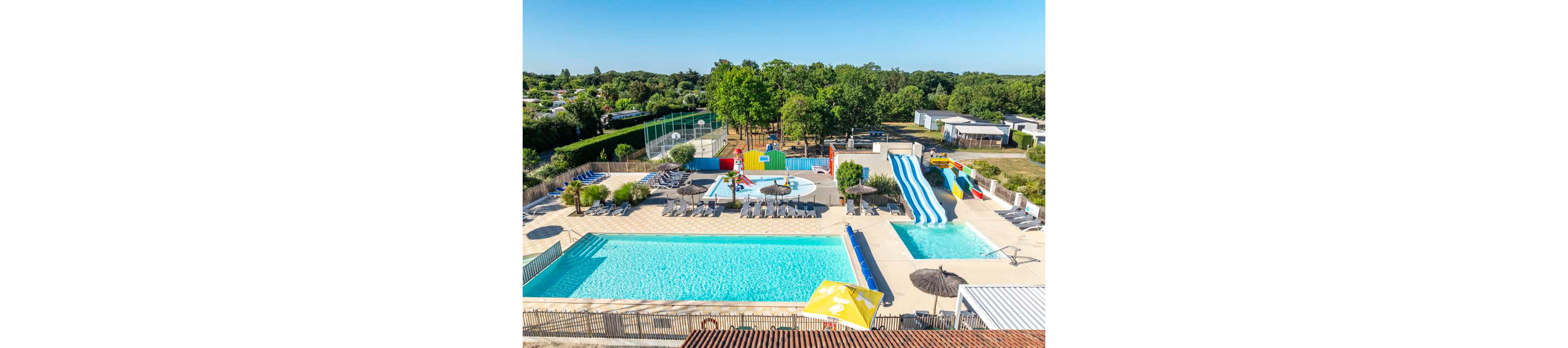 An aerial view shows an outdoor water park with multiple swimming pools and colorful water slides. A shallow splash area with small play structures sits between the main pools, surrounded by sun loungers and parasols. The complex is bordered by green trees, sports courts, and nearby cabins or mobile homes, suggesting a holiday resort setting. Bright blue water and clear skies create a sunny, inviting summer atmosphere.