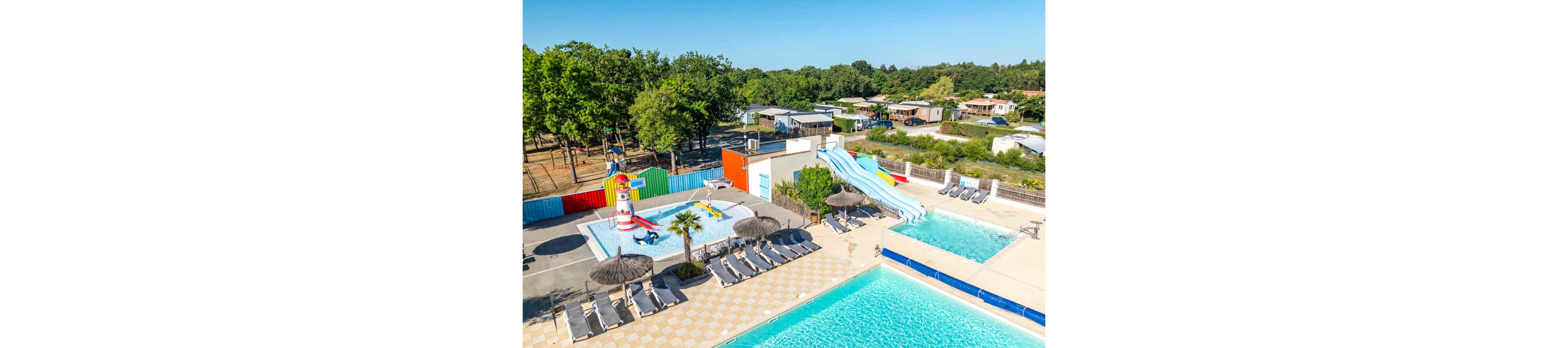 An outdoor campsite features multiple swimming pools and colorful water slides under a clear blue sky. A shallow splash area with playful fountains and a lighthouse-themed structure is designed for children. Sun loungers with straw parasols line the tiled pool deck, offering space for relaxation. In the background, mobile homes and trees create a peaceful holiday park setting.