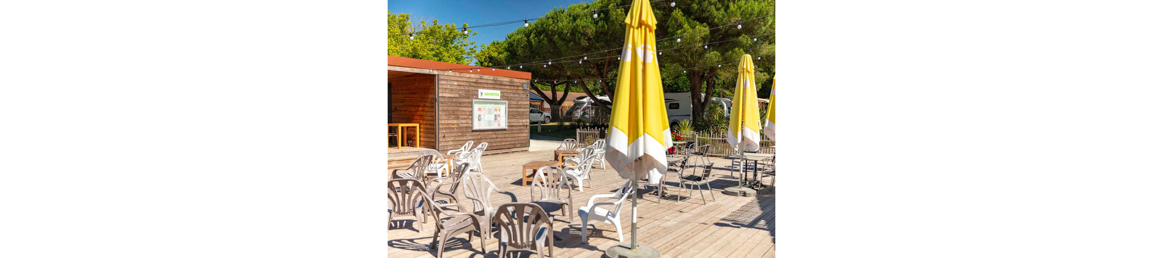 An outdoor café or bar terrace features numerous empty white plastic chairs arranged on a wooden deck. Large yellow and white umbrellas provide shade under a bright, sunny sky. A wooden kiosk or snack bar stands in the background with a menu board on its wall. String lights are hung overhead, and tall green trees surround the relaxed, holiday-like setting.