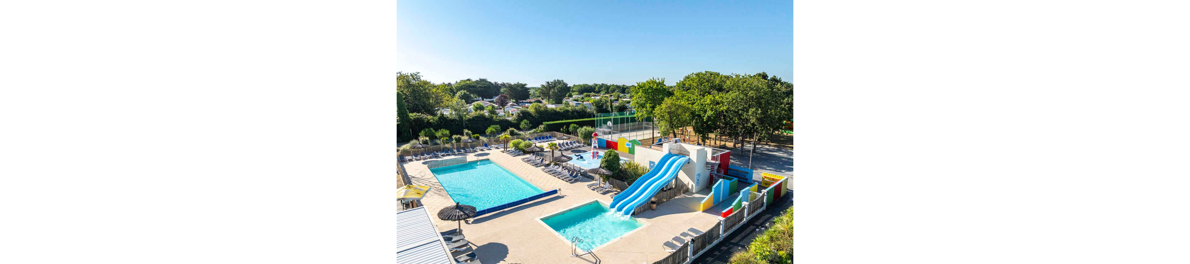 An outdoor aquatic center features a large rectangular swimming pool alongside a smaller shallow pool. A bright blue multi-lane waterslide curves into the water play area, surrounded by colorful structures and fencing. Sun loungers and parasols line the pool deck, with a lifeguard station visible near the center. Trees, greenery, and nearby recreational courts frame the scene under a clear blue sky, suggesting a sunny holiday resort atmosphere.