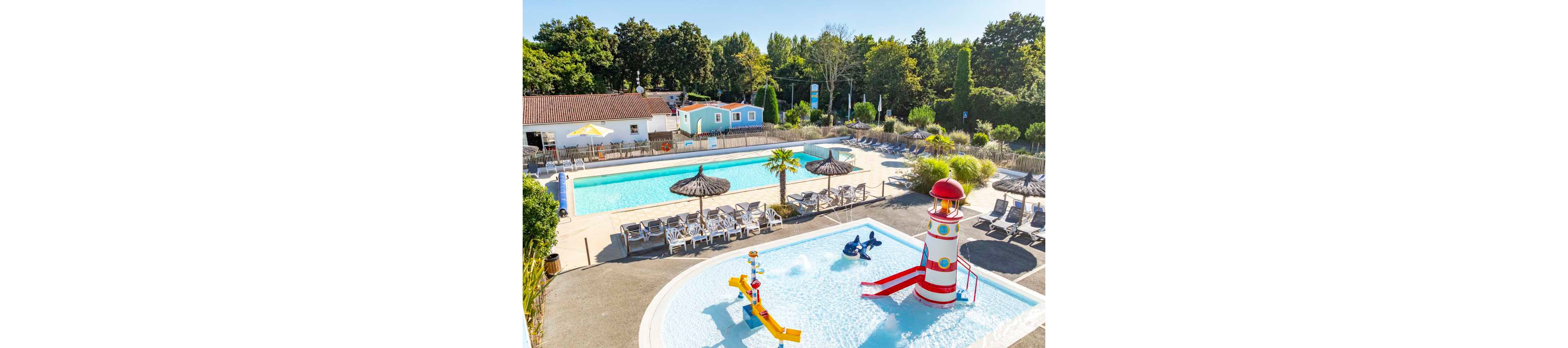 An outdoor holiday resort features multiple swimming pools surrounded by sun loungers and parasols. In the foreground, a shallow kids’ pool includes colorful water play structures and a lighthouse-themed slide. Behind it, a larger rectangular pool offers space for swimming and relaxation. Trees and small buildings frame the scene, creating a peaceful vacation atmosphere under clear skies.