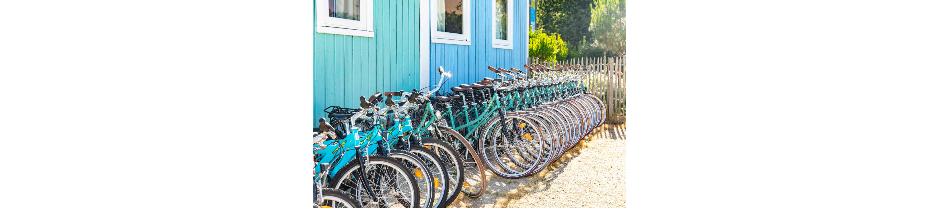 A long row of turquoise and black bicycles is neatly lined up outside a bright blue wooden building. The bikes are parked on a gravel path next to a rustic wooden fence. Sunlight creates strong highlights and shadows, suggesting a warm, clear day. The scene evokes a casual, summery atmosphere, typical of a vacation or coastal town bike rental spot.