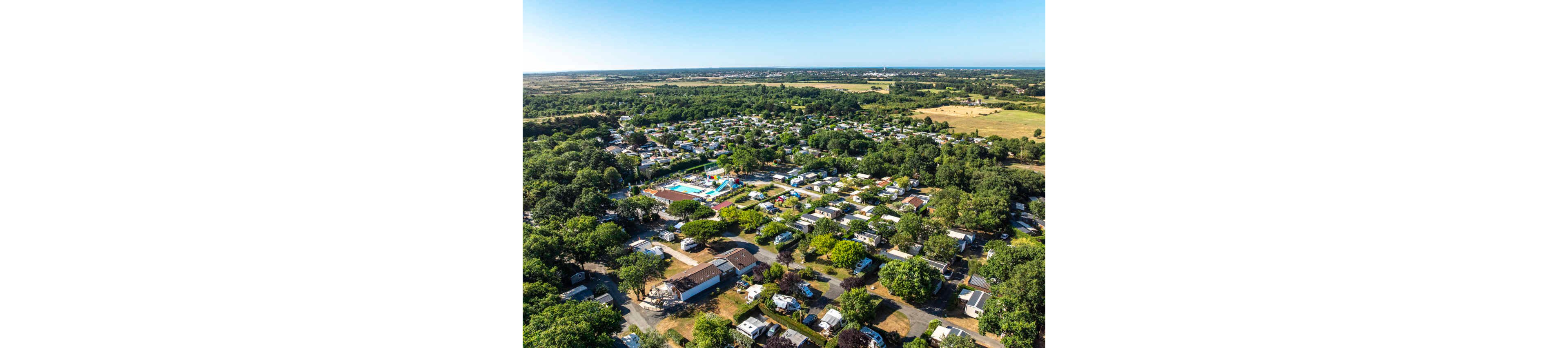 A wide aerial view shows a large campground or holiday park surrounded by trees and open countryside. Numerous white caravans, mobile homes, and small buildings are arranged in neat rows along internal roads. At the center, a colorful swimming pool and water play area stand out as a focal point. The surrounding landscape includes fields, patches of forest, and a distant horizon under a clear blue sky.