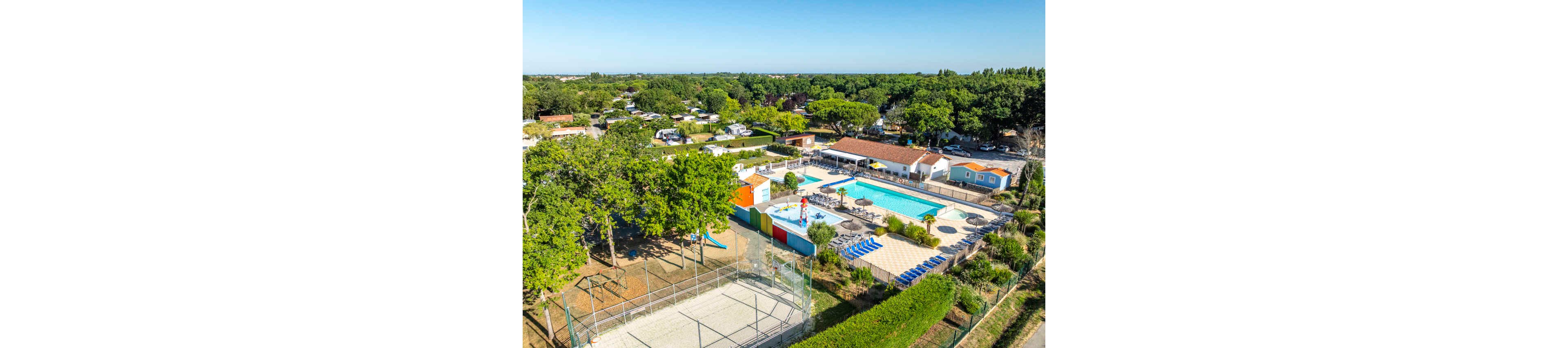 A large outdoor swimming pool complex is seen from above in a green, tree-filled campsite. The pool area includes a main pool, a smaller shallow pool with a waterslide, and rows of blue sun loungers. Nearby are low buildings with red-tiled roofs, colorful structures, and a fenced sports court. The surrounding area features caravans, mobile homes, and tents nestled among trees, creating a relaxed holiday atmosphere.