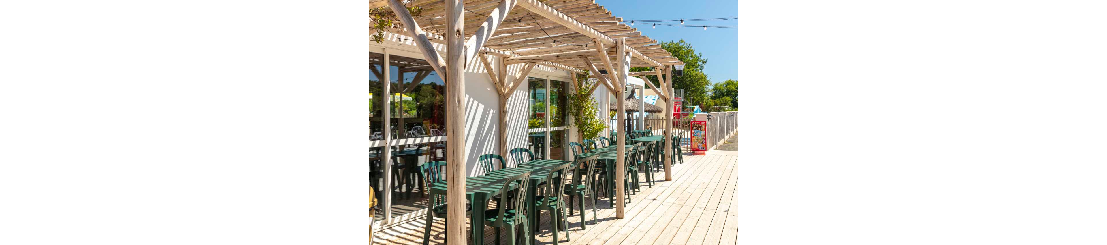 An empty outdoor café terrace features rows of green plastic tables and chairs beneath a light wooden pergola. The seating area is attached to a white building with large glass windows reflecting the bright day. String lights hang above the decked walkway, adding a festive touch to the scene. In the background, colorful vending or snack machines and lush greenery suggest a relaxed, summery atmosphere.
