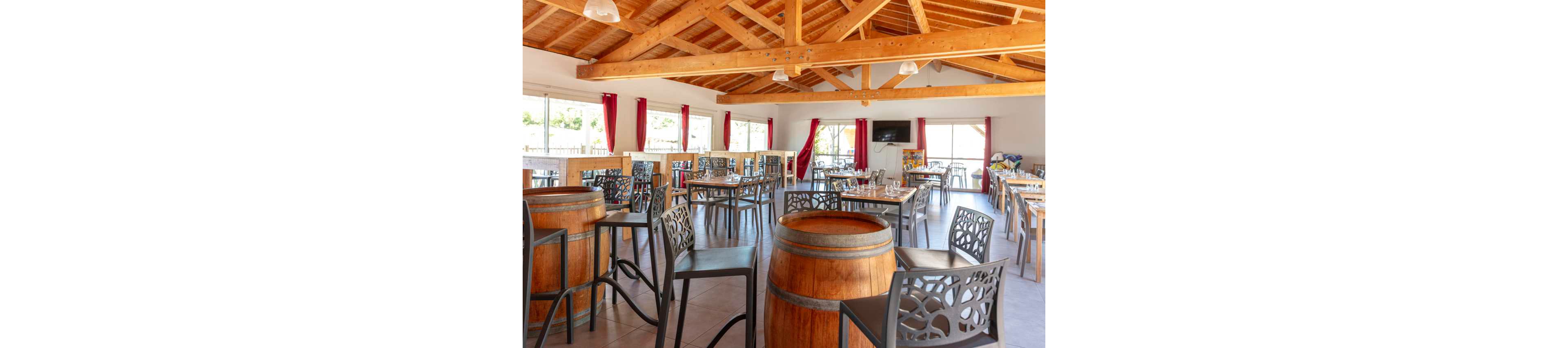 Spacious restaurant interior featuring wooden barrel tables and metal chairs under an exposed timber ceiling. Large windows with red curtains let in abundant natural light and offer views of the outdoors. Tables are set with glassware, cutlery, and napkins, ready to welcome guests. A wall-mounted television and neutral tiled floor complete the modern rustic dining atmosphere.