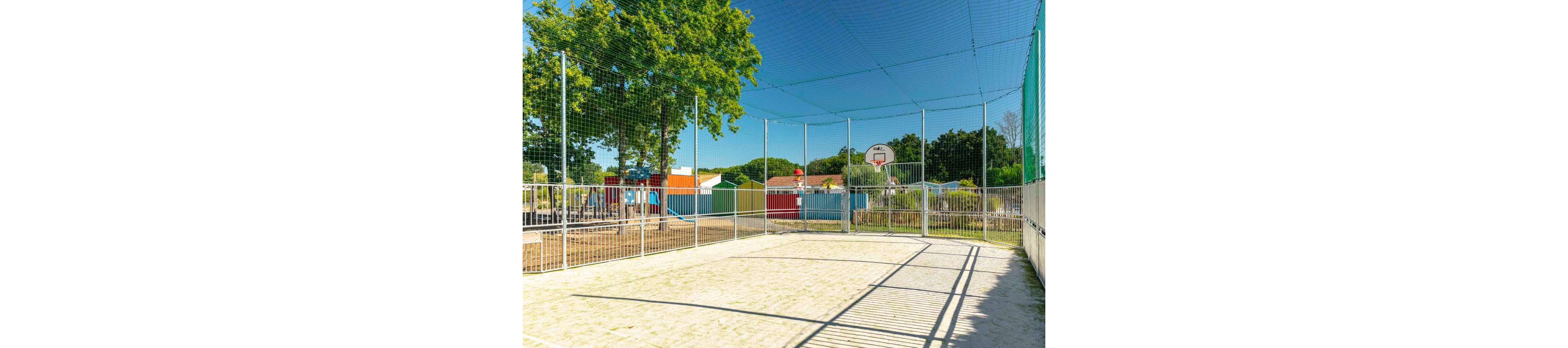 An empty outdoor sports court is enclosed by tall metal fencing and overhead netting. A single basketball hoop stands at one end of the court under a clear blue sky. In the background, colorful small buildings and a playground structure are visible among green trees. Bright sunlight casts long shadows across the sandy or gravel playing surface, creating a summery, recreational atmosphere.
