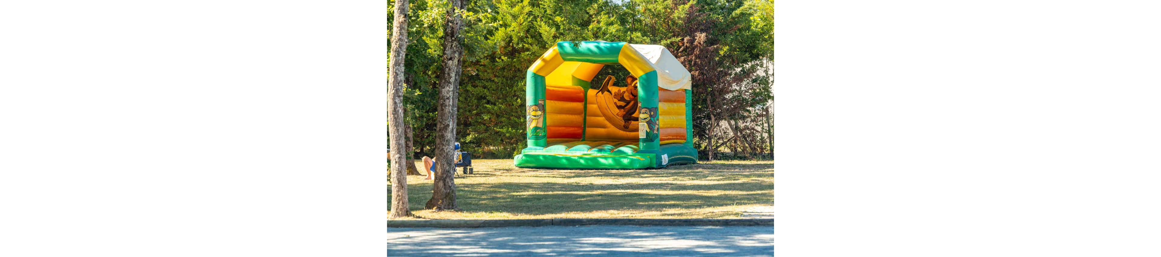 A colorful inflatable bounce house stands on a grassy area in a park. The structure features bright yellow, green, and orange panels with cartoon characters on the sides. Trees and bushes surround the play area, creating a natural backdrop. A child is visible nearby, enjoying the outdoor setting on a sunny day.