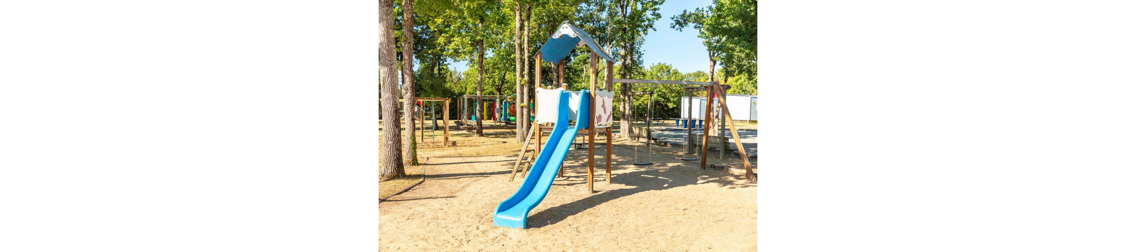 A bright blue slide stands in a sandy playground surrounded by tall trees. The play structure includes a small covered platform and wooden supports. Additional climbing frames and swings are visible in the background, indicating a larger play area. The scene is sunlit and quiet, with no children present.