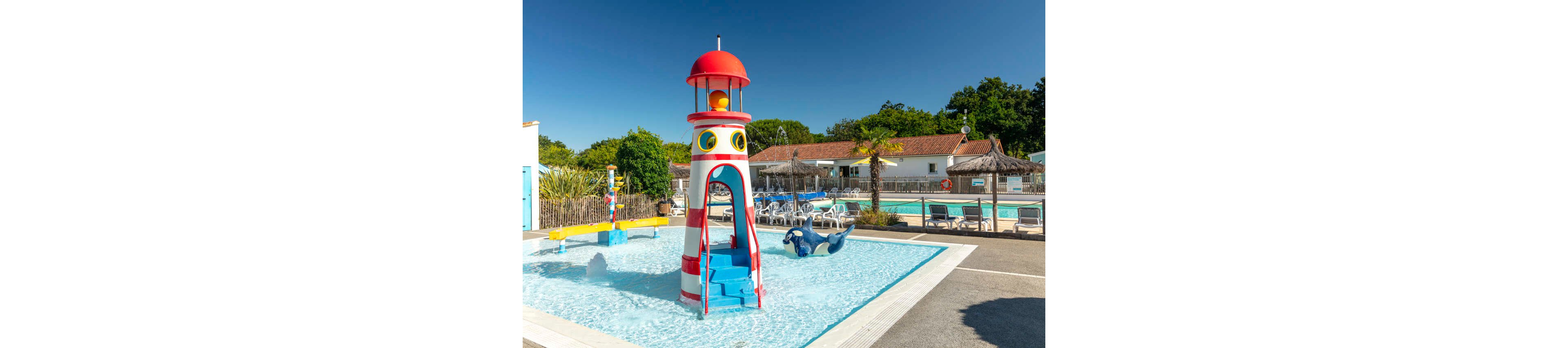 A colorful lighthouse-themed water play structure stands in a shallow splash pad pool. The scene is set at an outdoor resort or campground pool area with lounge chairs and umbrellas in the background. A playful dolphin slide and other water features add to the fun, family-friendly atmosphere. Bright sunlight and clear blue skies create a cheerful, summery mood.