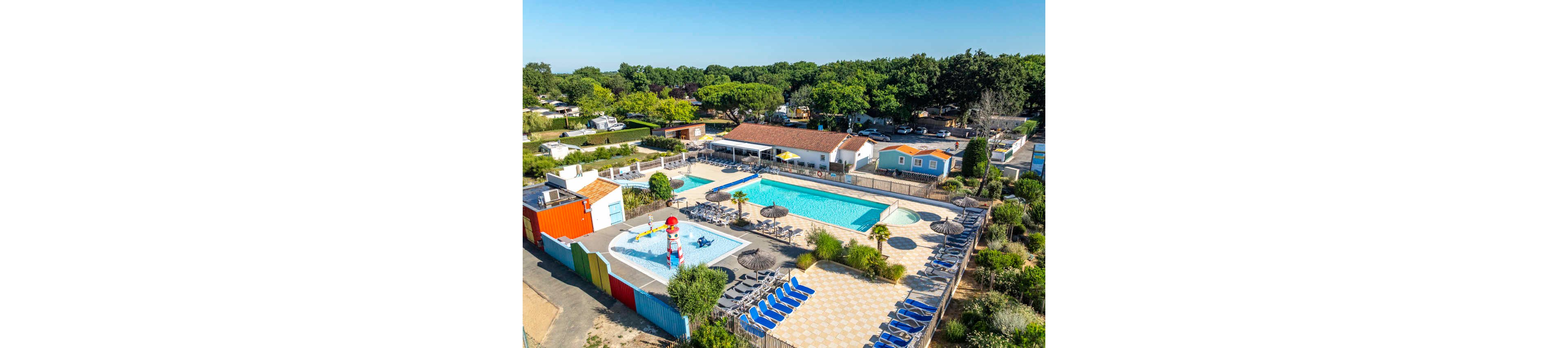 A large outdoor swimming pool complex is shown from above at a family campsite or holiday park. The scene includes a main rectangular pool, a smaller pool, and a colorful children’s splash area with a red-and-white lighthouse feature. Numerous blue sun loungers and parasols surround the pools, with landscaped greenery and pathways in between. In the background are small holiday cabins, caravans, and dense green trees under a clear blue sky.