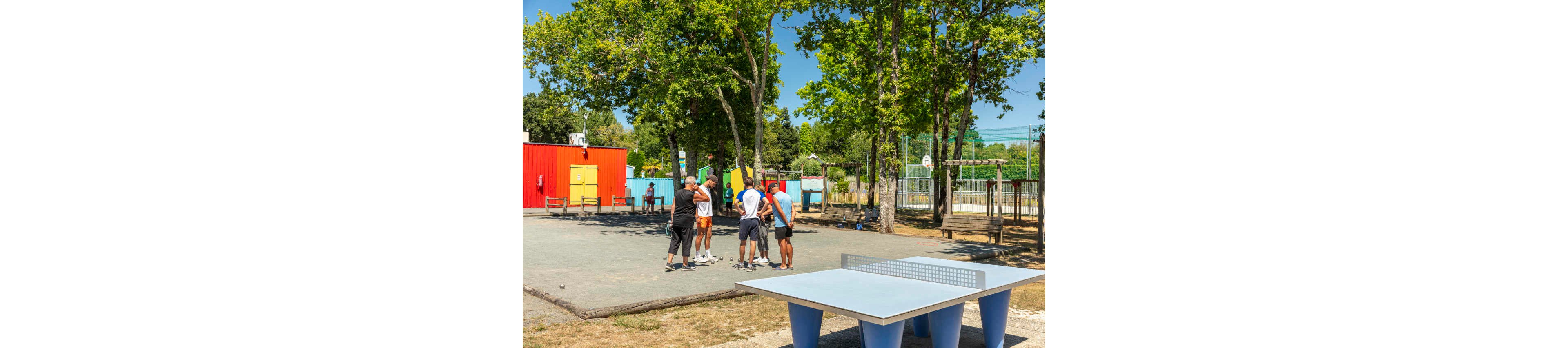 A small group of adults gathers on a gravel court to play pétanque in a sunny outdoor recreation area. They stand in a circle discussing the game while trees provide shade around them. In the foreground, an empty outdoor table tennis table is visible, and in the background there are colorful buildings, benches, and a fenced basketball court. The scene suggests a relaxed summer day focused on leisure and social activity.