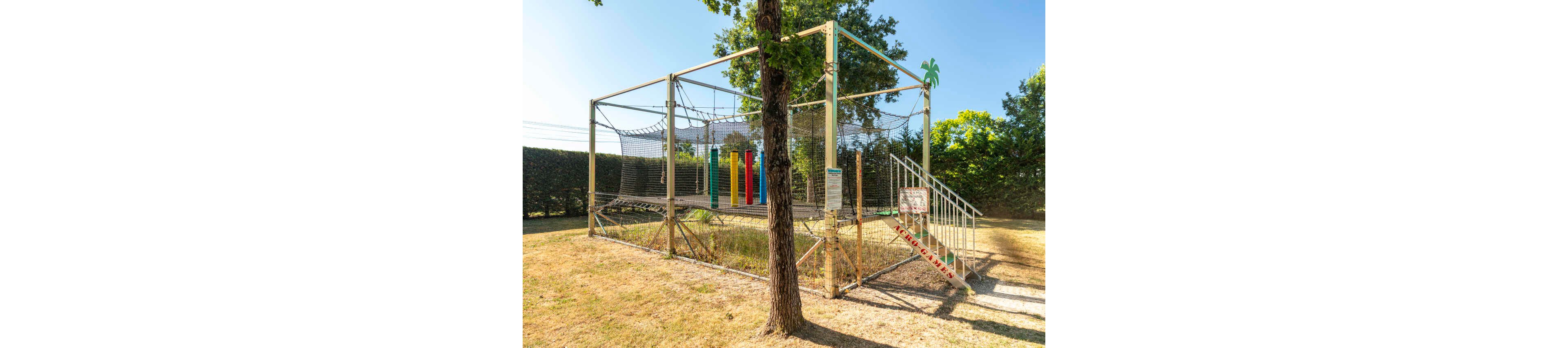 An elevated outdoor trampoline play area is enclosed with safety nets and metal framing in a grassy yard. Colorful vertical foam pillars stand inside the structure, suggesting a children’s activity zone. A metal staircase with handrails leads up to the platform, and a tree trunk stands in the foreground partially obscuring the view. Hedges and trees surround the area, creating a sunny, park-like setting.