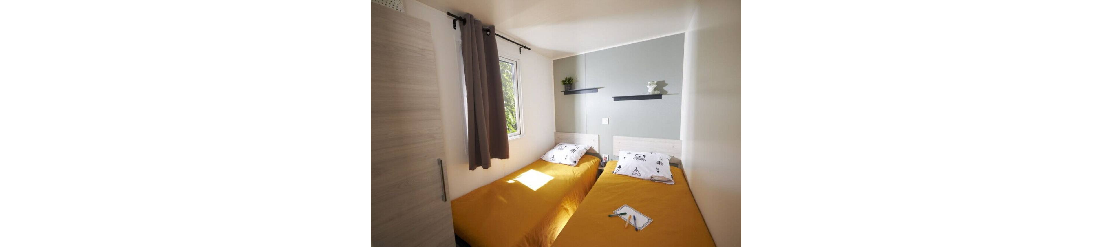 Compact twin bedroom featuring two single beds with bright yellow duvets and patterned pillows. Natural light enters through a small window with gray curtains, casting sunlight onto the beds. A gray accent wall holds two minimalist shelves with small decorative items. The room has a clean, modern style with light wood furniture and neutral tones.