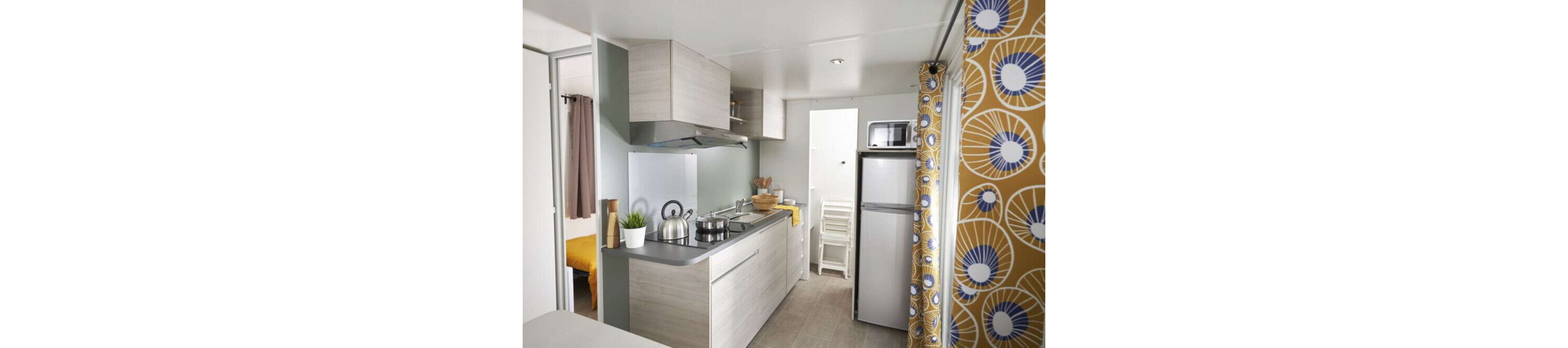 A compact modern kitchen features light wood cabinets, a slim countertop, and stainless steel appliances. A kettle and saucepan sit on the stovetop beside a small potted plant and wooden utensils. A tall refrigerator with a microwave on top stands near stacked white chairs in the background. A doorway reveals a cozy bedroom with yellow bedding, while a bold patterned curtain adds color to the bright interior.