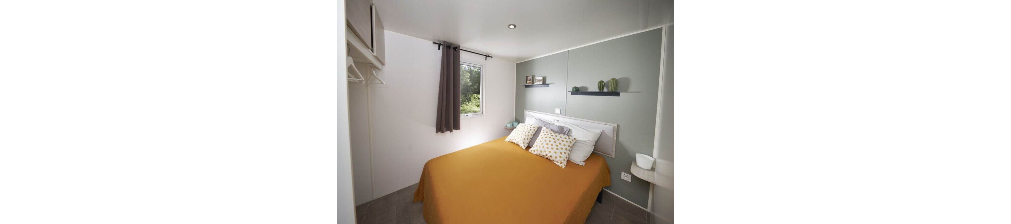 A small modern bedroom features a double bed with a mustard-colored bedspread and patterned pillows. The room has light walls with a muted green accent panel behind the headboard. A window with gray curtains lets in natural light and offers a view of greenery outside. Minimalist shelves with decor items and a small side table complete the simple, contemporary design.
