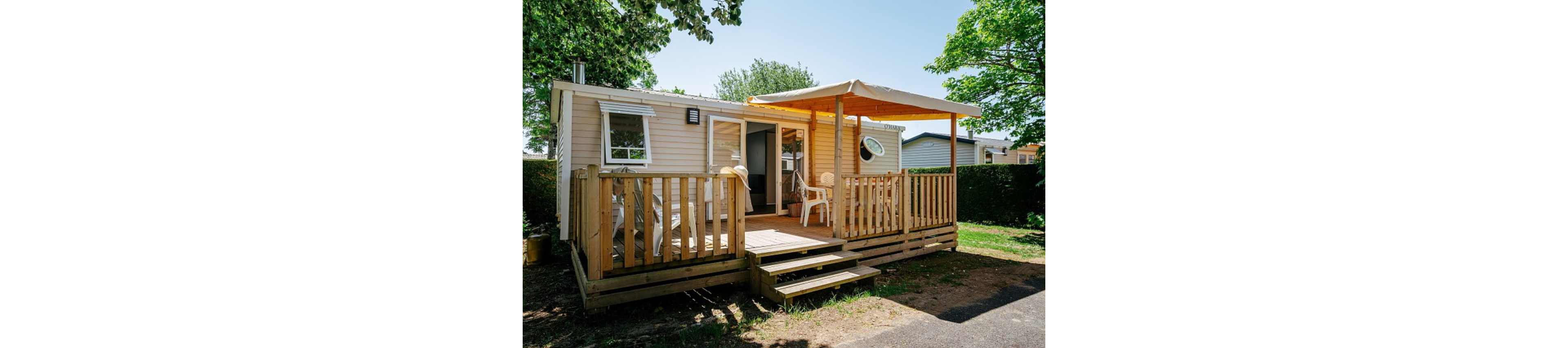 A compact mobile home features a wooden deck with railing and steps leading to the entrance. The terrace is partially covered by a canopy, providing shade for white outdoor chairs and a small table. Surrounded by green trees and hedges, the home sits in a quiet, sunny campground setting. The scene suggests a relaxed vacation atmosphere in a modest, comfortable accommodation.