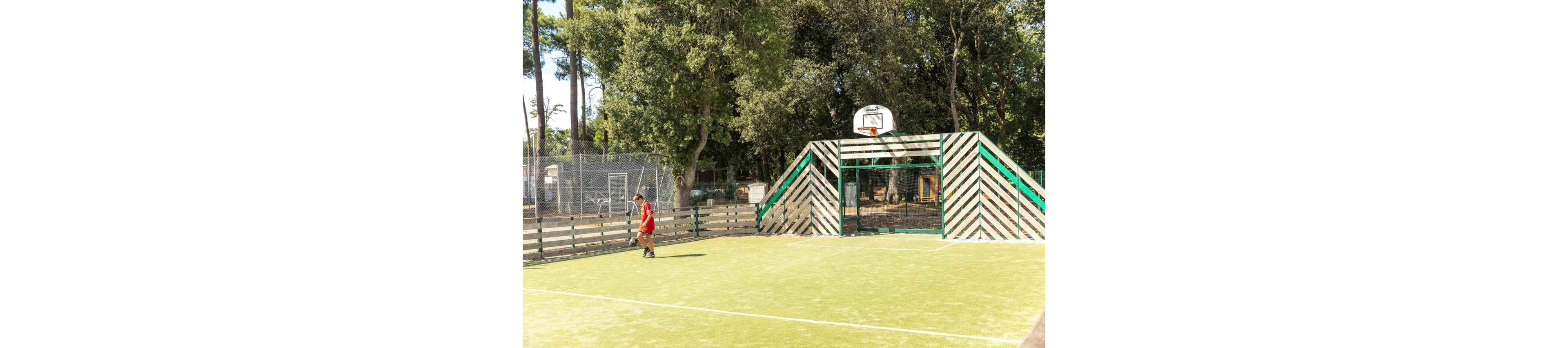 An outdoor sports court features a small soccer goal combined with a basketball hoop above it. A person in a red shirt walks along the edge of the green artificial turf. Wooden fencing and striped wooden structures frame the goal area. Tall trees and a wire fence surround the court, creating a sunny park-like setting.
