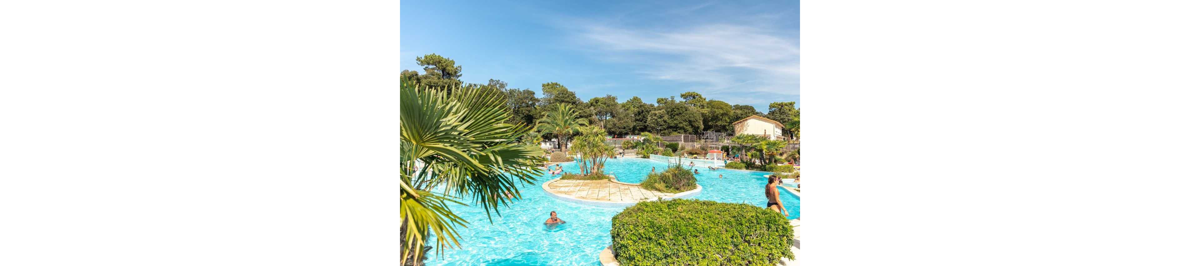 A large outdoor swimming pool winds through a landscaped resort area surrounded by lush greenery and palm trees. Several people relax and swim in the clear blue water under a bright, sunny sky. Low buildings and dense trees frame the background, giving the scene a secluded holiday atmosphere. The overall setting suggests a warm-weather vacation destination focused on leisure and family fun.