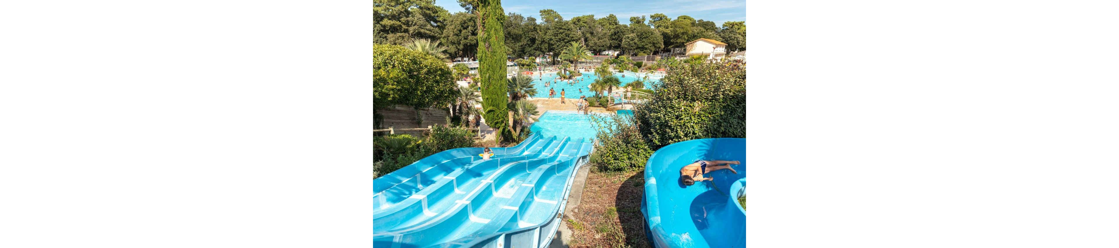 An outdoor water park features multiple bright blue water slides leading into a large swimming pool. Families and children enjoy the sunny day, swimming and playing in the clear blue water. Lush green trees, shrubs, and palm trees surround the pool area, creating a resort-like atmosphere. The scene captures an energetic summer holiday mood with people relaxing and having fun in the water.