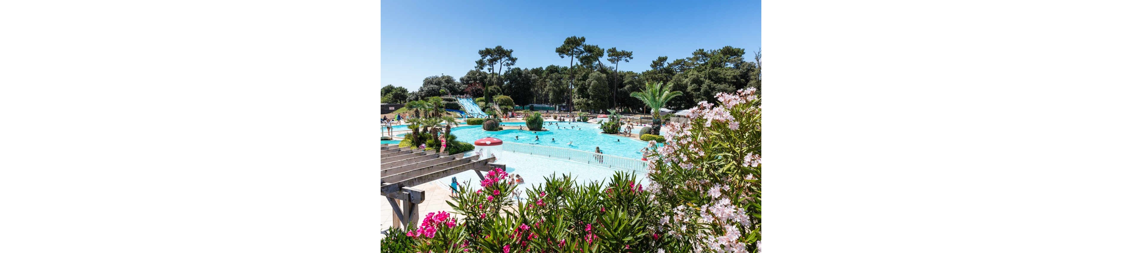 An outdoor swimming pool is surrounded by lush greenery and flowering bushes on a bright sunny day. Families and children enjoy swimming and playing in the water, with several people visible in the pool. In the background, tall trees frame the scene and multiple blue waterslides lead into the pool area. The clear blue sky and vibrant plants create a cheerful, summery atmosphere.