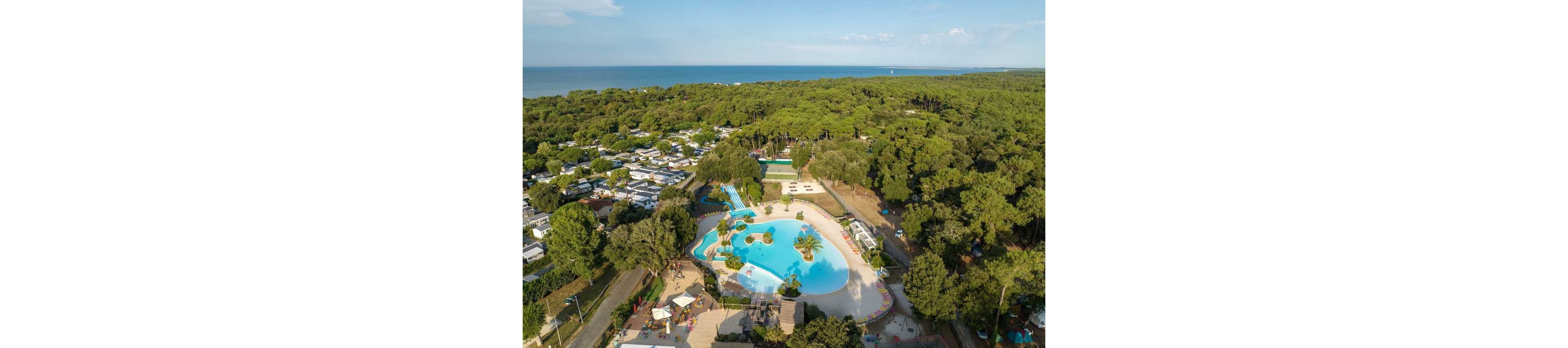 A large outdoor swimming pool complex is nestled among dense green forest near a coastline. The aerial view shows winding blue pools with small islands, water slides, and surrounding sun loungers. Nearby, numerous white mobile homes or cabins are arranged in neat rows under the trees. In the distance, the calm sea stretches toward the horizon under a clear sky.