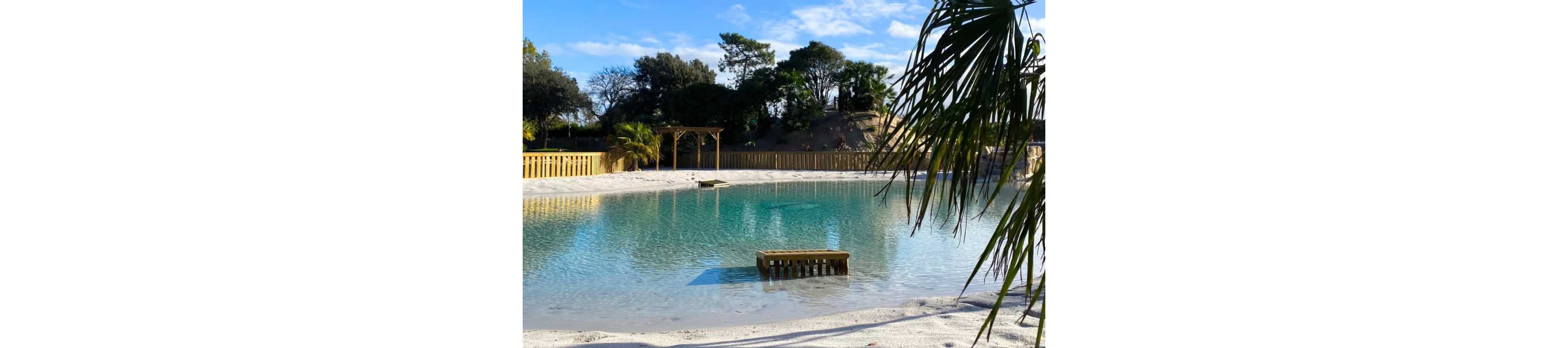 A tranquil turquoise lagoon-style pool sits in a sandy area surrounded by wooden fencing. A small wooden platform extends into the water, reflecting the clear blue sky above. Palm trees and lush greenery frame the scene, giving it a tropical, resort-like atmosphere. In the background, a wooden pergola and distant trees complete the peaceful outdoor setting.