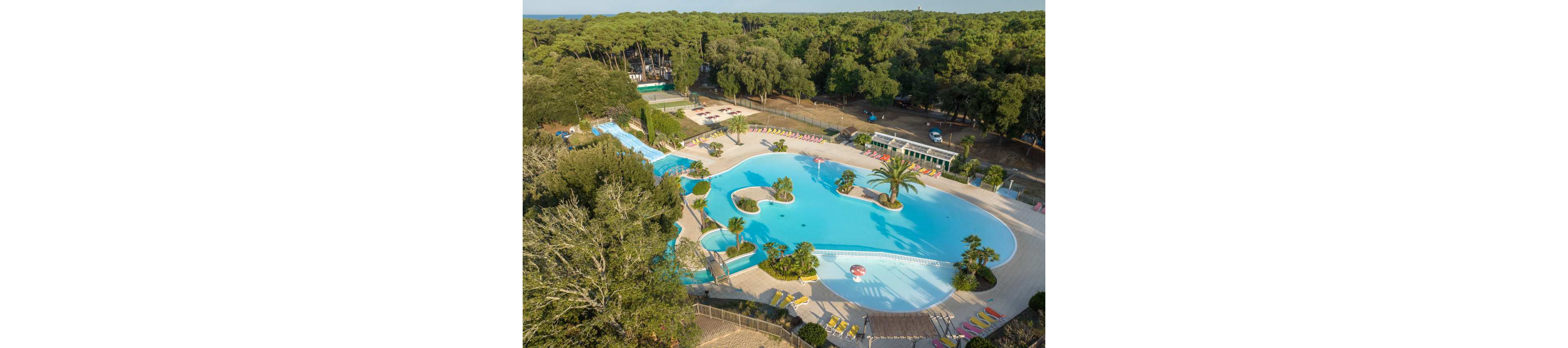 A large, lagoon-style swimming pool is surrounded by sun loungers and palm trees in a forested resort setting. The pool features curved edges, shallow areas, and a small island with a tall palm tree. Nearby, additional pools and recreational areas extend into the trees, with tennis courts visible in the background. Dense green woodland encircles the entire complex, creating a secluded holiday atmosphere.