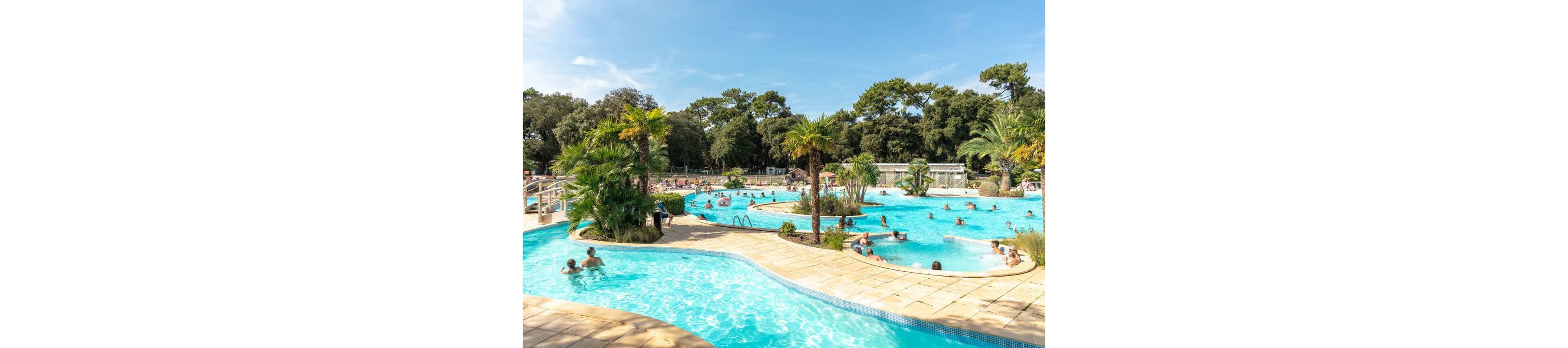 An expansive outdoor swimming pool complex features winding turquoise pools surrounded by palm trees and greenery. Families and couples relax in the water and on the sun loungers along the tiled pool deck. The scene is bright and sunny with clear blue skies, creating a summery vacation atmosphere. Buildings and additional facilities are visible in the background, suggesting a holiday resort setting.