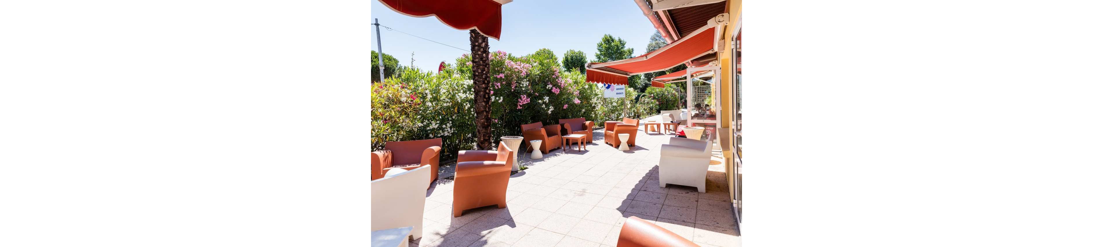 An outdoor cafe terrace features modern plastic armchairs arranged on a tiled patio. Red awnings cast partial shade while bright sunlight creates strong shadows across the ground. Lush green bushes and flowering plants form a colorful backdrop around the seating area. The scene suggests a relaxed, summery atmosphere ideal for socializing or taking a break outside.
