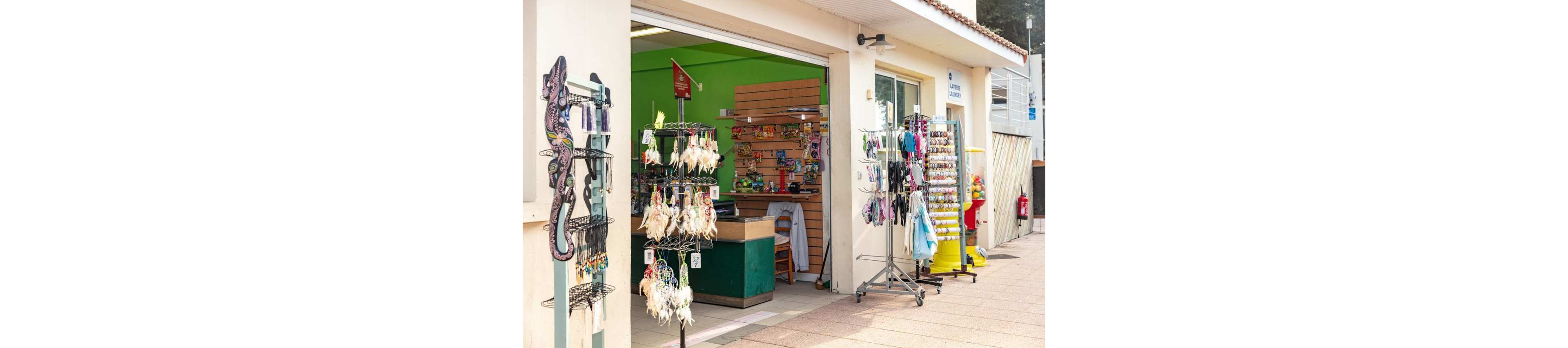 An open-front souvenir shop displays various accessories and trinkets on racks outside the entrance. Colorful keychains, dreamcatchers, and small decorative items hang from metal stands on the sidewalk. Inside the shop, shelves and a counter are visible against a bright green wall. The scene suggests a casual tourist location on a sunny day.