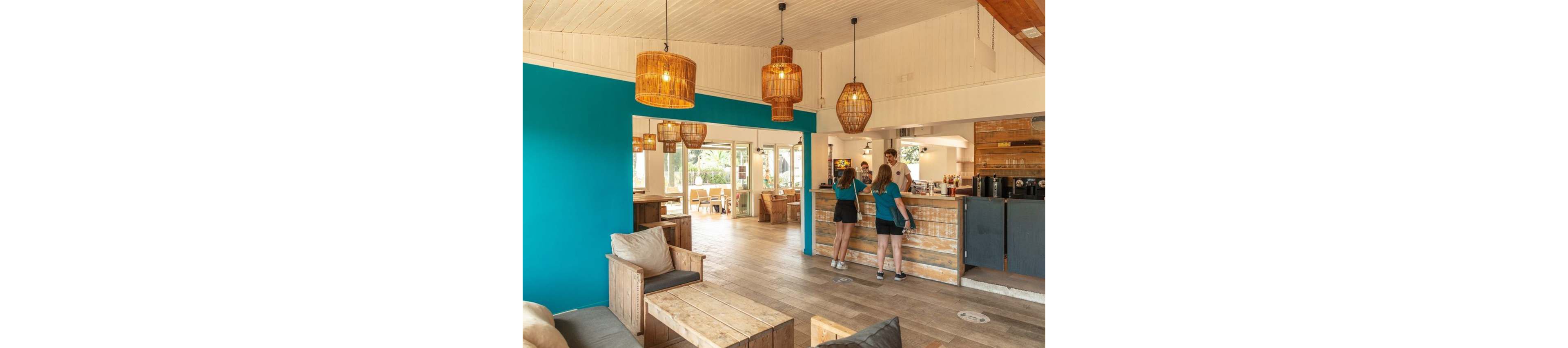 A bright cafe interior features wooden furniture and a turquoise accent wall. Two young women stand at the counter, speaking with a staff member in a relaxed setting. Large wicker pendant lights hang from the white ceiling, casting a warm glow over the space. Sunlight streams through glass doors at the back, revealing an outdoor seating area and greenery.