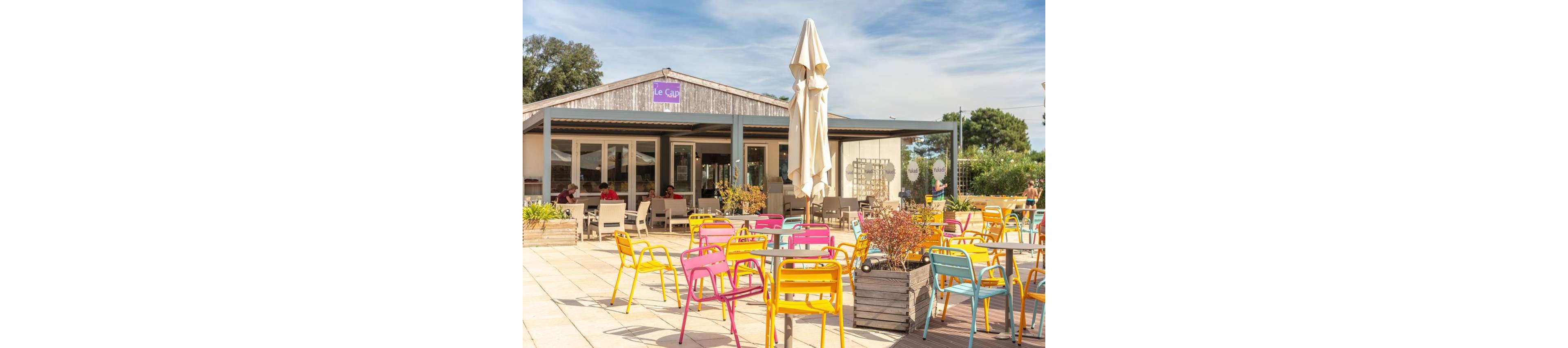An outdoor café terrace features numerous brightly colored metal chairs and tables under a sunny sky. The café building in the background has a wooden facade with large windows and a visible sign reading "Le Café." A few people sit near the entrance, enjoying the relaxed atmosphere. Potted plants and greenery surround the seating area, adding to the casual, summery feel of the scene.