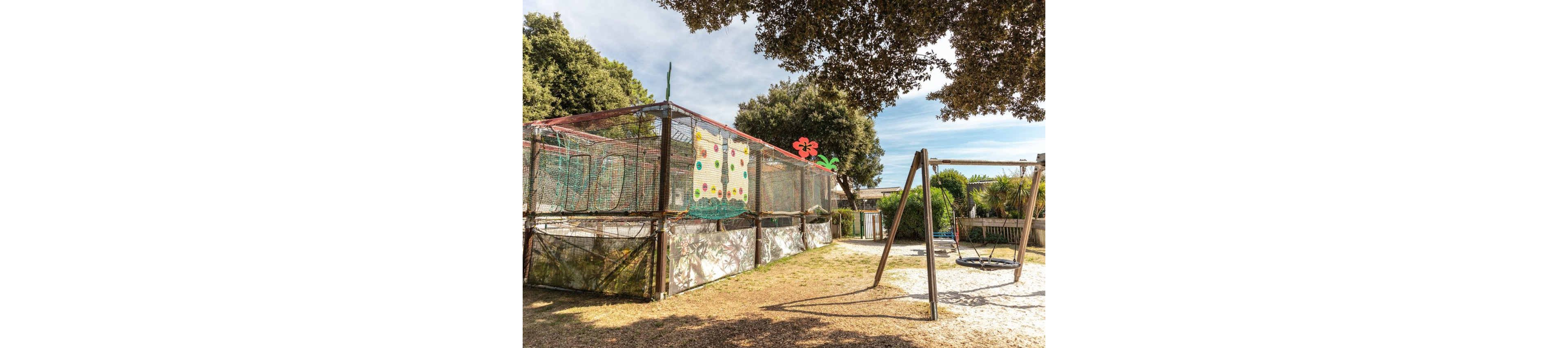 An outdoor playground features a large netted play court decorated with colorful panels and a red flower illustration. Beside it stands a metal swing set with a circular swing over a sandy surface. Trees and shrubs surround the area, providing shade and a natural backdrop. The scene is bright and sunny, suggesting a pleasant day for children’s outdoor activities.