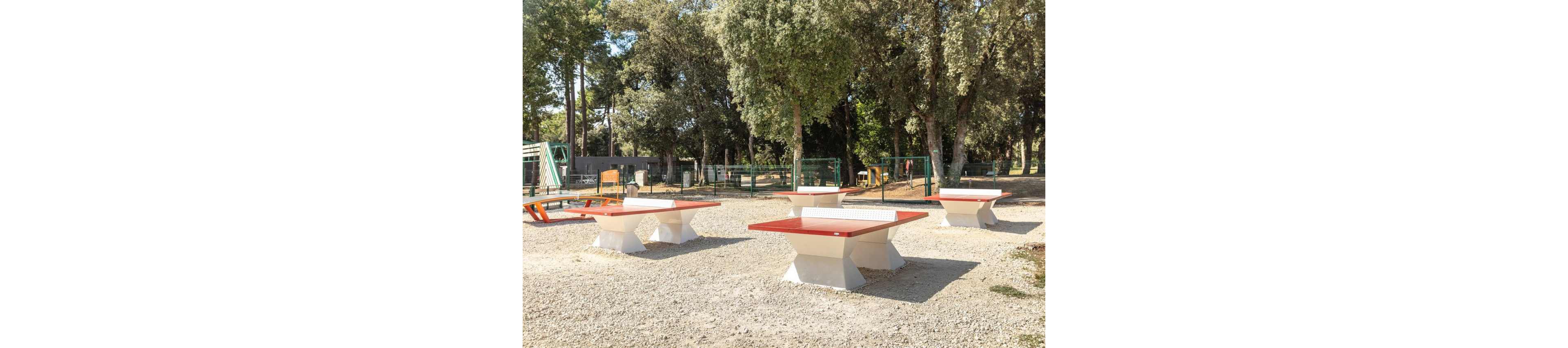 Several red and white concrete ping pong tables stand in an open gravel area. The setting appears to be an outdoor recreation zone bordered by green metal fencing and surrounded by tall trees. Benches and playground-style structures are visible in the background, suggesting a family-friendly park. Bright daylight and clear shadows create a warm, inviting atmosphere for outdoor games.