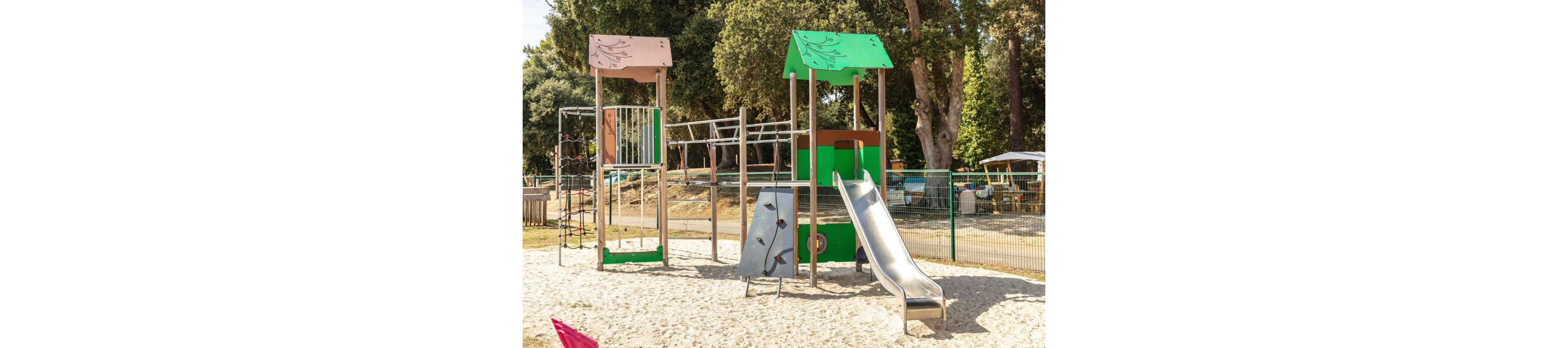 An empty outdoor playground stands on a sandy surface under bright daylight. The play structure features a tall metal slide, climbing wall, rope net, and monkey bars between two colorful towers. Green trees and a wire fence surround the play area, with park seating and vehicles visible in the background. The scene suggests a family-friendly recreational space in a public park or campground.