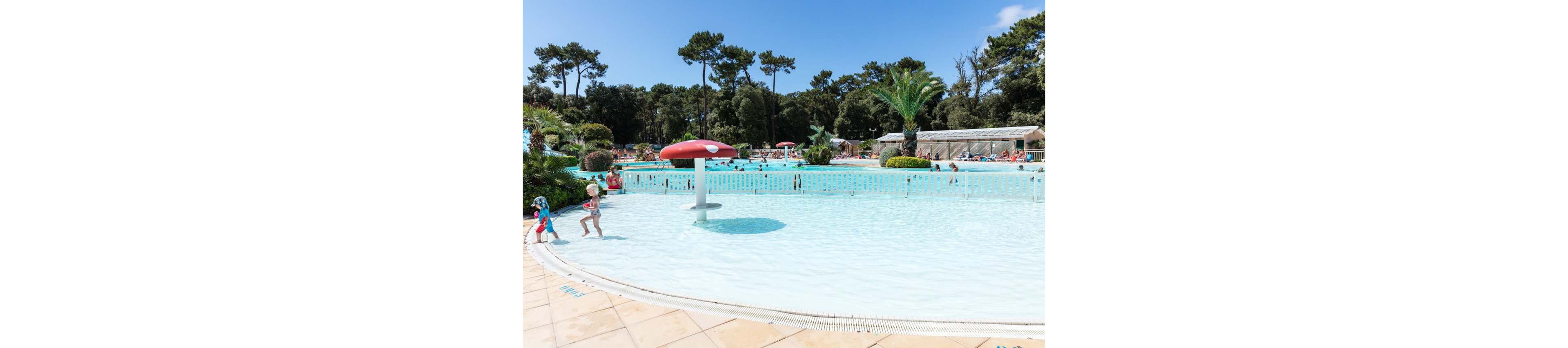 A shallow outdoor swimming pool features young children playing near a red mushroom-shaped water fountain. The pool is surrounded by a tiled deck and a white safety fence. In the background, more people relax and swim in additional pools under a clear blue sky. Tall pine trees and lush greenery frame the family-friendly water park setting.
