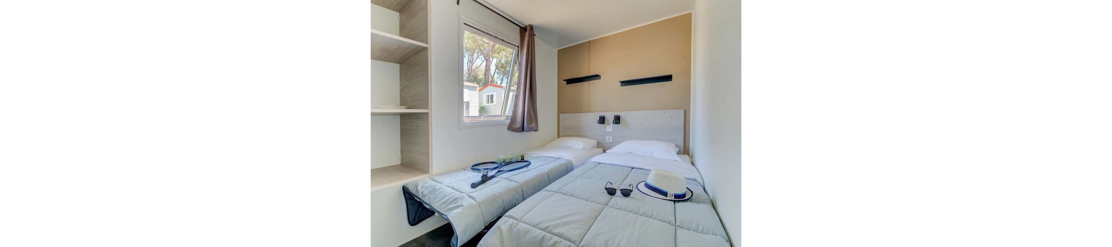 Compact bedroom with two single beds is shown in a modern vacation cabin. A window with a brown curtain lets in daylight and reveals neighboring mobile homes and trees outside. One bed holds a tennis racket and ball, while the other has folded towels and sunglasses, suggesting a relaxed holiday stay. Built-in shelves, neutral walls, and simple wall lamps create a clean, minimalist atmosphere.
