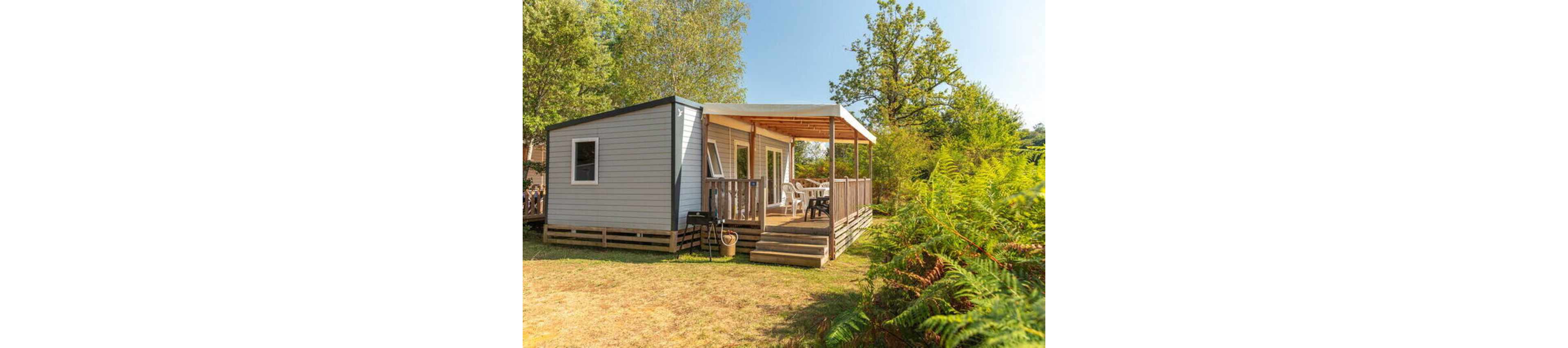 A modern mobile home stands in a sunny outdoor setting surrounded by trees and greenery. The structure features light-colored siding, several windows, and a covered wooden terrace with steps leading down to the grass. Plastic chairs and a table are arranged on the deck, suggesting a relaxed vacation or camping atmosphere. The scene is bright and inviting, with warm sunlight and lush foliage creating a peaceful holiday mood.