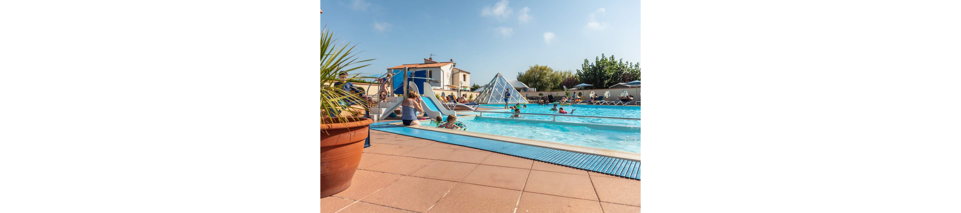 Children and adults relax and play at a bright outdoor swimming pool. A colorful inflatable ring sits on the poolside tiles in the foreground, while kids use water slides and splash in the water. The setting appears to be a family-friendly resort or holiday park with sun loungers and umbrellas. Clear blue sky and warm sunlight create a cheerful, summery atmosphere.