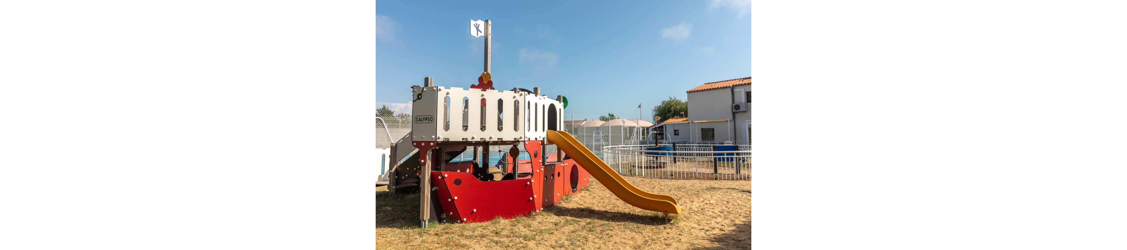A colorful pirate ship themed playground structure stands on a sandy play area. Two children are visible playing inside the structure, which features a bright yellow slide and red lower panels. A small white flag flies at the top of the mast, and the word "CALYPSO" appears on the side. Residential-style buildings and a fenced yard surround the playground under a clear blue sky.