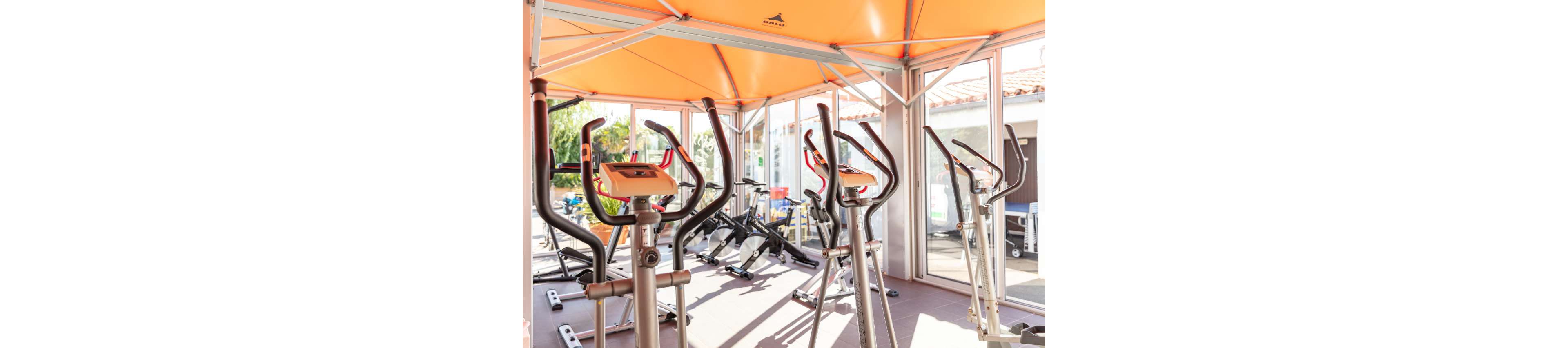 A sunlit indoor gym features several elliptical trainers in the foreground. Stationary bikes and additional fitness equipment are visible in the background. Large glass windows and doors let in abundant natural light, creating an open and airy atmosphere. An orange ceiling canopy adds a vibrant accent to the modern workout space.