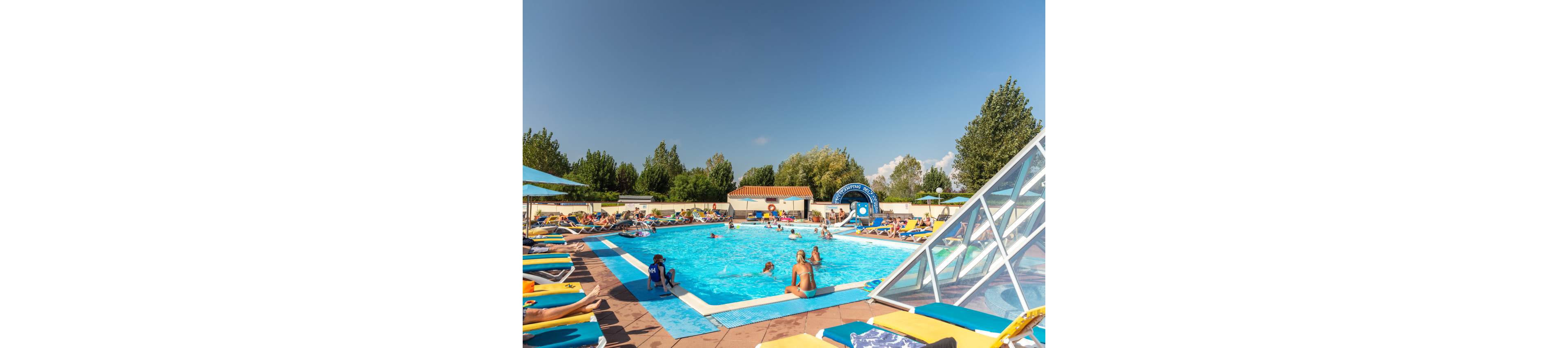 An outdoor swimming pool is filled with people relaxing and playing in the water on a sunny day. Sun loungers with colorful towels and umbrellas surround the pool, where families and children enjoy the holiday atmosphere. A small slide and water feature are visible near the far end, while trees and a clear blue sky frame the scene. The overall mood is lively, warm, and summery.