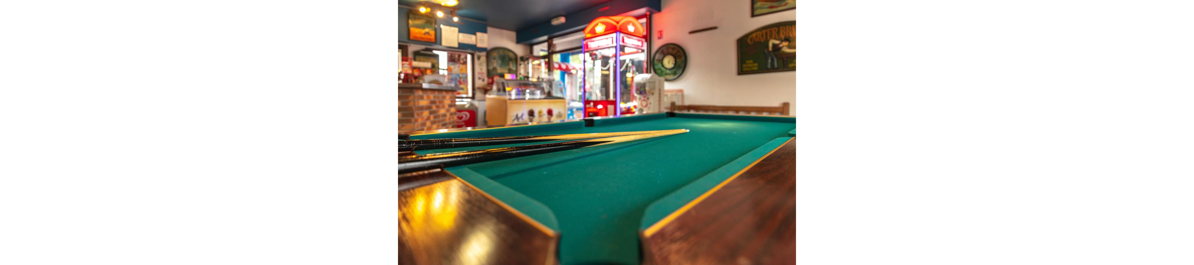 A green felt pool table with several cues rests in the foreground of a lively bar or game room. In the background, a brightly lit arcade claw machine and snack counter add color and activity. Warm lighting and vivid reds, greens, and yellows create a fun, casual atmosphere. Posters and wall decor contribute to the relaxed entertainment setting.
