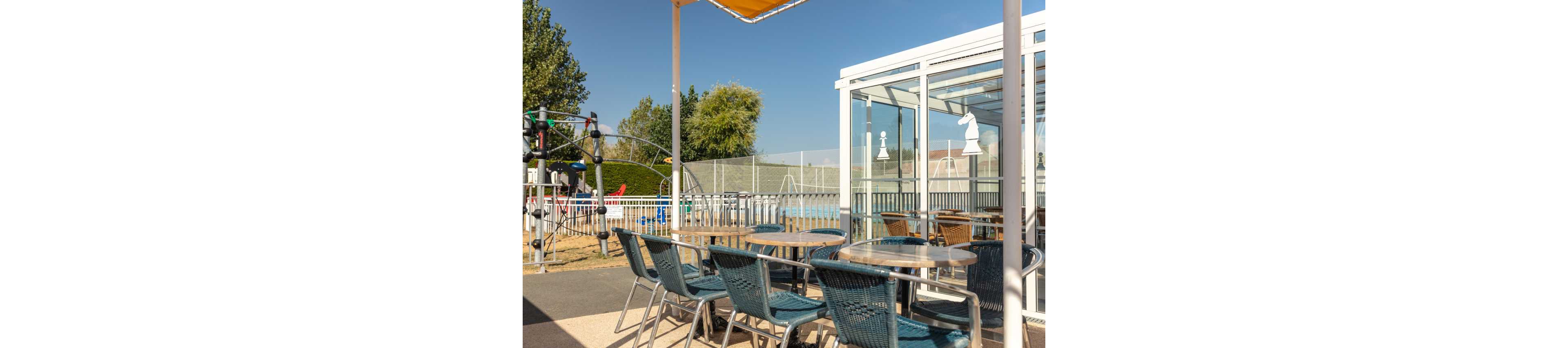 An outdoor café terrace features several metal tables with woven chairs under a yellow awning. Beyond the terrace, a fenced swimming pool and a children's playground with climbing structures are visible. A glass-enclosed seating area with reflections and chess piece decals stands to the right. The scene is brightly lit by clear blue sky, suggesting a warm, sunny day in a leisure or resort setting.